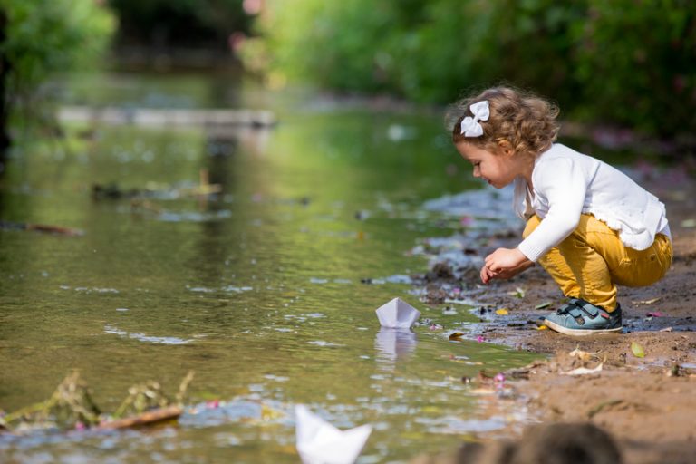 Cute little girl runs a paper boat in the stream in the park. Stretching her hand and reaching the little ship