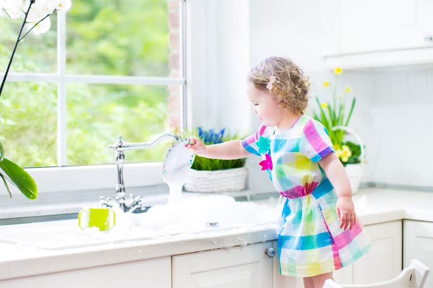 Cute curly toddler girl in a colorful dress washing dishes, clea
