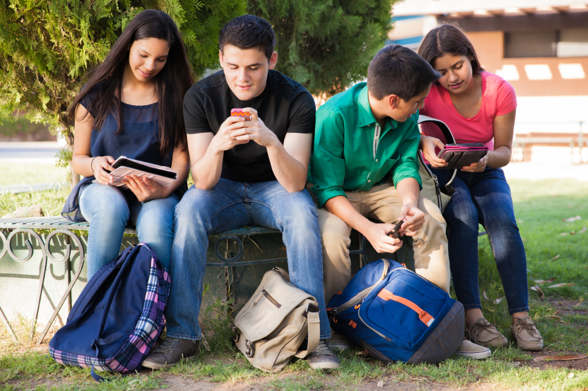 Group of high school students using cell phones and tablets while relaxing outdoors